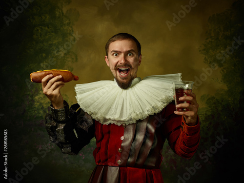 Young man as a medieval knight on dark studio background. Portrait of male model in retro costume. Holding a glass of beer and hot-dog. Human emotions, comparison of eras, facial expressions concept.