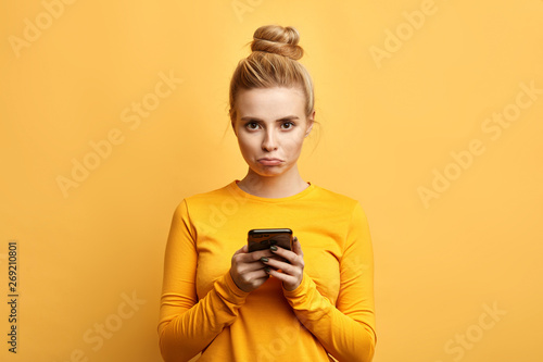 angry sad woman with hairbun annoyed by something while using phone, girl has received bad sms, text message isolated yellow background. emotion, reaction, feeling