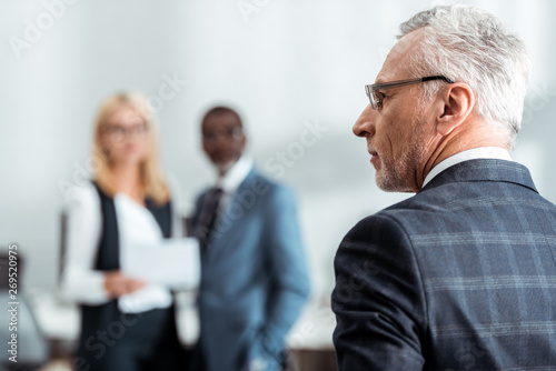 selective focus of man in glasses near multicultural colleagues