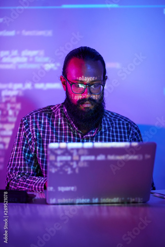 Bearded programmer in eyeglasses concentrating on his work, he sitting at the table working on laptop in dark office