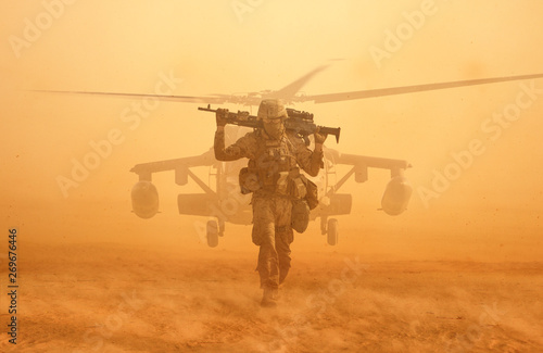 Military soldier walking at desert with gun on his shoulder in front of helicopter in sand storm.