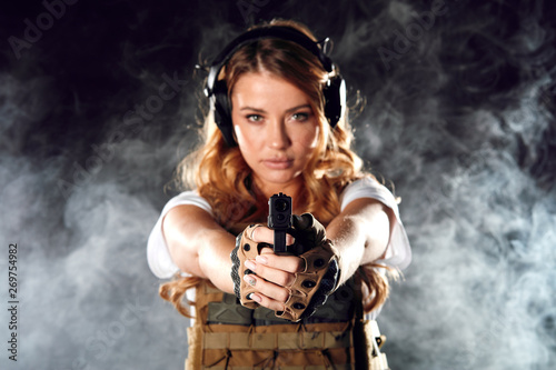 Armed beautiful blonde woman wearing protective headphones and plate carrier, shoots with gun at a target in the darkness with smoke clouds