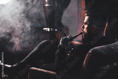 Groomed bearded man is relaxing on lounge near fireplace while smoking hookah. He has tattoo on his hand.