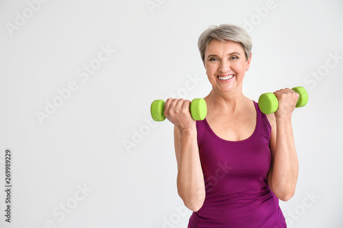 Sporty mature woman with dumbbells on light background