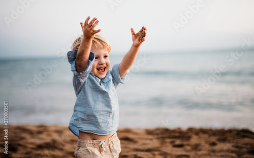 A small toddler boy standing on beach on summer holiday, having fun.