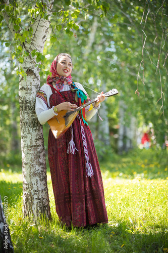 Young romantic woman in traditional russian clothes standing in the forest and holding balalaika