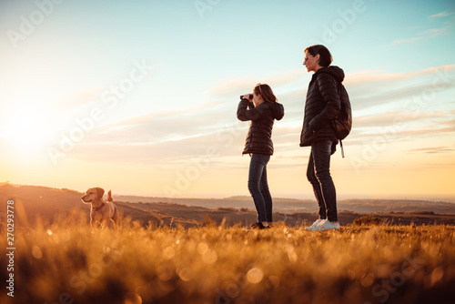 Mother and daughter standing on top of mountain with dog and using binoculars