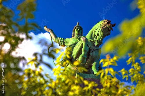 Majestic bronze green monument to Salawat Yulaev, close-up shot through spring foliage on a bright summer day. Folk hero on a horse with whip in his hand. Salavat Yulayev, Ufa, Bashkortostan, Russia.