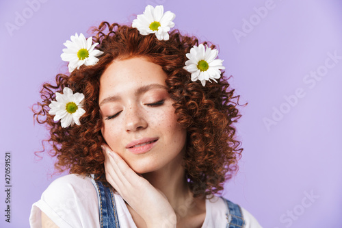 Beautiful curly happy redhead girl posing isolated over purple background with flowers in hair.