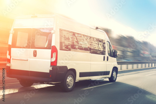 Unrecognizable small passenger van hurry up on highway at city street traffic with urban cityscape and sunset sky on background. Charter or shuttle bus van hurry up on road with motion blur effect