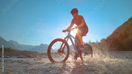 LOW ANGLE: Fit man rides an e-bike in the shallow river and splashes water.