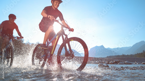 LOW ANGLE: Water drops fly around two guys riding electric bicycles along river.