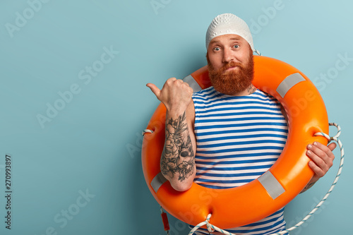 Photo of puzzled male swimmer or instructor points thumb left, attracts your attention to copy space, wears swimhat and striped vest, holds ring buoy, isolated on blue background. Advertisement