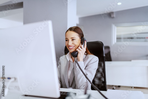 Gorgeous Caucasian businesswoman with long brown hair and in formal wear using computer and talking on the phone. Multitasking concept.