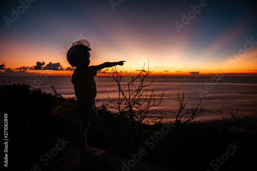 A boy in a helmet looks at the sunset at sea on the viewpoint