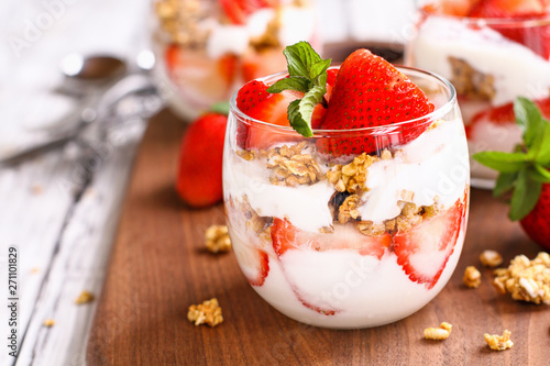 Healthy breakfast of strawberry parfaits made with fresh fruit, yogurt and granola over a rustic white table. Shallow depth of field with selective focus on glass jar in front. Blurred background.