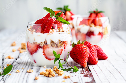 Healthy breakfast of strawberry parfaits made with fresh fruit, yogurt and granola over a rustic white table. Shallow depth of field with selective focus on glass jar in front. Blurred background.