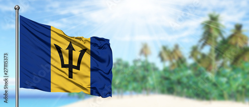Waving Barbados flag in the sunny blue sky with summer beach background. Vacation theme, holiday concept.