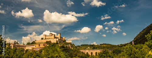 View of Spoleto, green mountains, blue sky with white clouds. The Rocca Albornoziana fortress illuminated by the sun in summer. The bridge of the towers, Roman aqueduct. Trees in the foreground