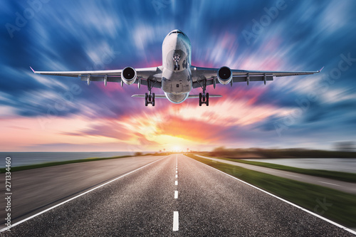 Airplane and road with motion blur effect at sunset. Landscape with passenger airplane is flying over asphalt road and colorful sky. Commercial plane is landing. Aircraft with blurred background