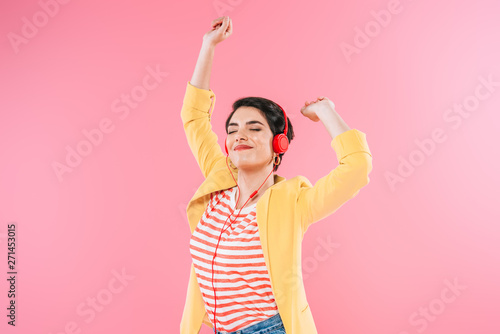 excited mixed race woman listening music in headphones and dancing isolated on pink