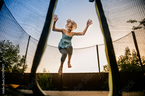 Child girl jumping high on big trampoline outside in garden on summer sunset