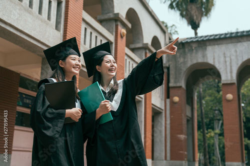 two young girls college students on graduation day holding textbook study abroad finish university. woman point finger showing friend smiling laughing talking chatting in traditional red brick house.