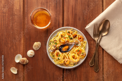 Seafood pasta. Tagliolini with mussels, shrimps, clams and squid rings, with a glass of wine, shells, a fork, and a spoon, shot from above on a dark rustic wooden background with copy space