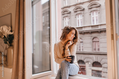 Romantic female model in good mood looking at street, sitting on sill. Fascinating dark-haired lady spending morning at home, chilling near window with coffee.