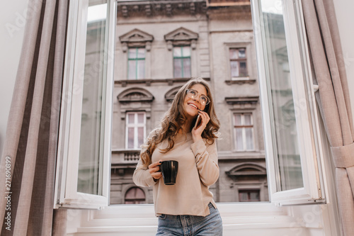 Elegant curly woman in glasses standing with cup of tea on urban background. Dreamy brunette girl in beige shirt talking on phone in front of opened window.