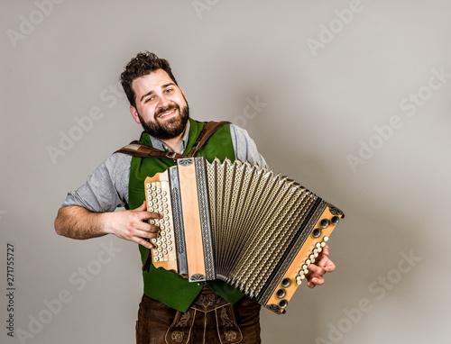 cool young musician with black beard and leather trousers and traditional costume and accordion is posing in front of grey background