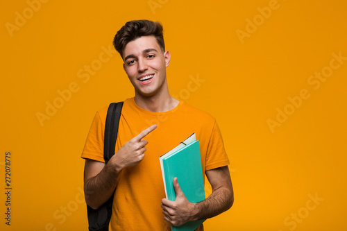 Young student man holding books pointing with finger at you as if inviting come closer.