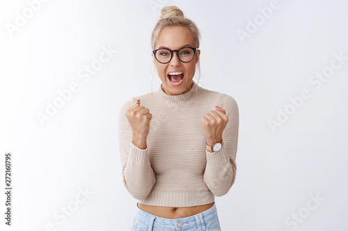 Yes we did it. Happy cheerful surprised beautiful glamour blond woman in glasses celebrating great awesome news clenching fists triumphing cheering and yelling from happiness over white wall