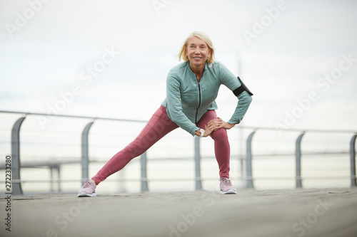 Happy mature sportswoman in activewear enjoying workout in urban environment while doing stretching exercise