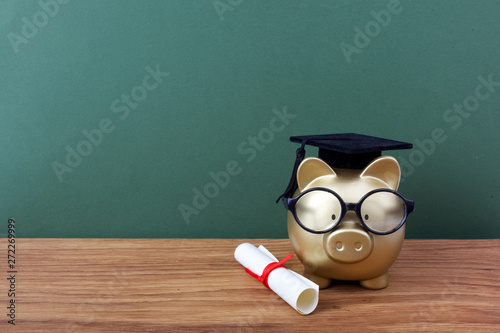 Gfold piggy bank with a grad cap and diploma in front of green chalkboard. Education scholarship