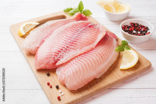 Raw fish fillet of tilapia on a cutting Board with lemon and spices. White table with copy space.