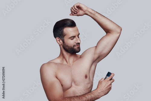 Freshness. Handsome young man applying deodorant and smiling while standing against grey background