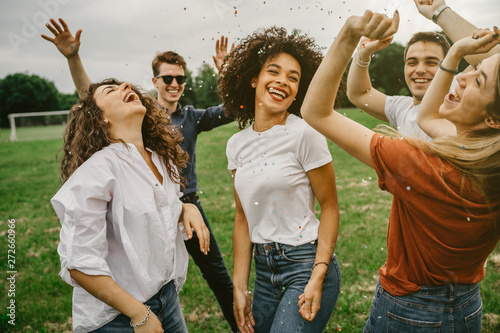 Group of five friends having fun at the park - Millennials dancing in a meadow among confetti thrown in the air - Day of freedom and carefree