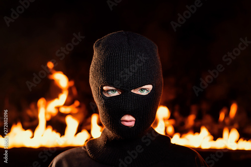 Portrait of young woman in black balaclava against backdrop of a blazing night fire. Concept of mass rallies and riots