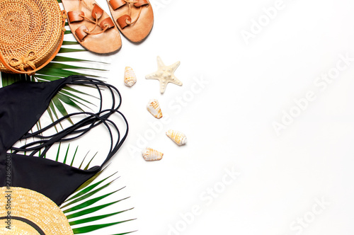 Woman's beach accessories flat lay. Round trendy rattan bag straw hat black swimsuit leather sandals tropical palm leaves seashells on white background. Top view copy space. Summer backdrop