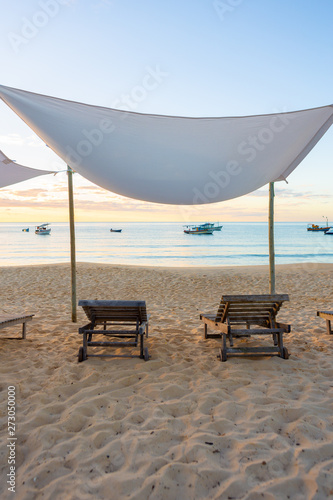 Beautiful beach with wooden chairs, sun loungers and tent with boats and sunset in the background.Concept of vacations, peace and relaxation. Ponta do Corumbau, Bahia, Brazil.