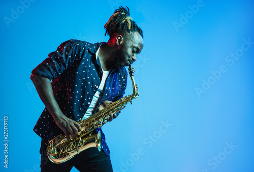 Young african-american jazz musician playing the saxophone on blue studio background in trendy neon light. Concept of music, hobby. Joyful attractive guy improvising. Retro colorful portrait of artist