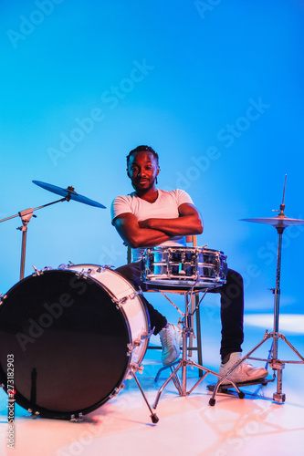 Young african-american jazz musician or drummer playing drums on blue studio background in trendy neon lights. Concept of music, hobby, inspirness. Colorful portrait of joyful attractive artist.