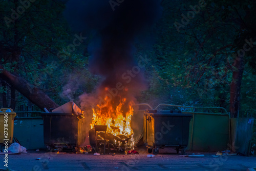 Burning trash container and old wood chair with other garbage. Plastic garbage can has almost fused