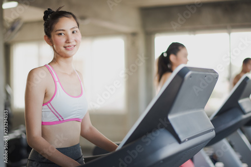 Young Asian woman walking and running on the running machine and looking camera at the gym. Woman workout running at fitness room. People training at gym concept.