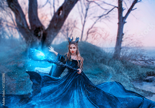 strict witch with magical magical glowing and sparkling staff in her hands, lady with horns on head utters spell, emerald silk flying long dress, demonessa on grassy bank of river during sunset