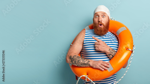 Studio shot of stupefied lifeguard points away on left side, keeps mouth opened from amazement, dressed in casual swimwear, poses with lifebuoy, isolated on blue background, free space for your advert