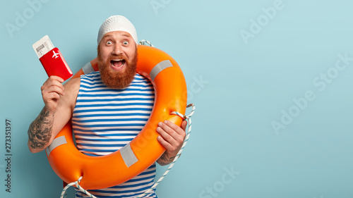 Happy joyous male vacationist enjoys summer time and holiday, ready for flight abroad, holds passport and flying tickets, wears swimhat and sailor vest, gazes with excitement. Vacation concept