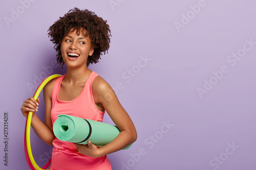 Fitness woman with dark skin, works with hula hoops, holds rolled karemat for training, laughs joyfully, satisfied after workout, looks healthy and fit wears rosy t shirt isolated on purple background