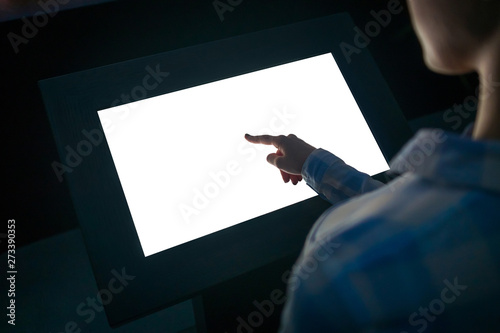 Woman hand using white blank interactive touchscreen display of electronic multimedia kiosk in dark room - scrolling and touching - close up view. Mock up, copyspace, template and technology concept